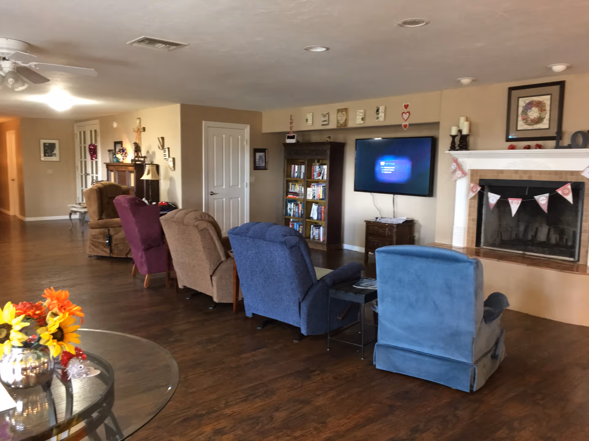 A cozy living room with several upholstered recliner chairs arranged in a row facing a wall-mounted flat screen TV. There is a fireplace decorated with a garland and candles on the mantel. A bookshelf filled with books is next to the TV. The room has wooden flooring, beige walls, and a ceiling fan. A glass table with a vase of colorful flowers is visible in the foreground.