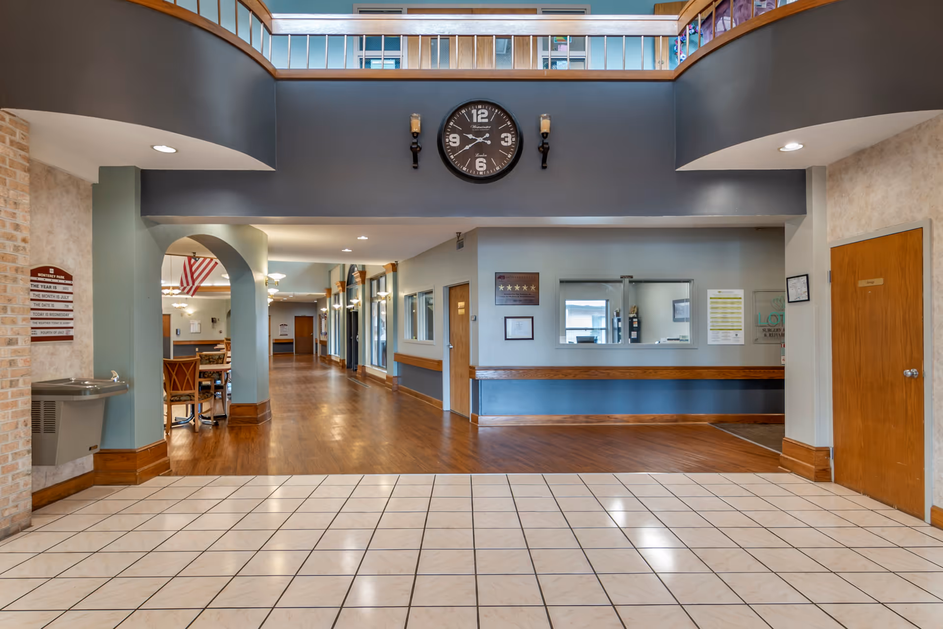 Interior view of a senior living facility hallway with a tiled floor in the foreground and wooden flooring further inside. There is a large clock mounted on a dark blue wall above an open reception window. To the left, there is a drinking fountain and a sign with the date and weather information. The hallway has wooden trim and doors, with seating and tables visible in the background under an American flag.