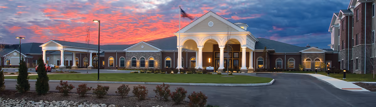 Exterior view of St Clare Commons senior living facility at dusk, featuring a large building with a prominent entrance supported by white columns, an American flag flying above, well-maintained landscaping, and a driveway leading to the entrance under a colorful sunset sky.