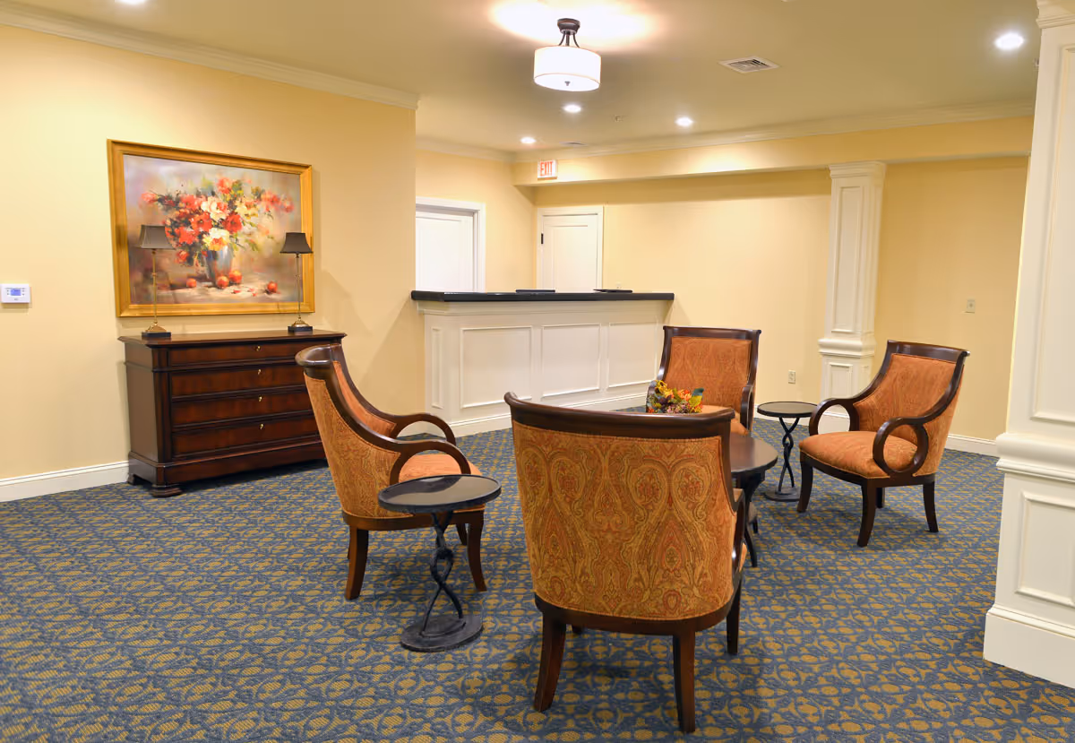 A cozy seating area in an assisted living facility with four upholstered armchairs arranged around a small round table with a floral centerpiece. The room has patterned blue and gold carpet, cream-colored walls, a wooden chest of drawers with two lamps and a large floral painting above it, and a white reception desk in the background under ceiling lights.