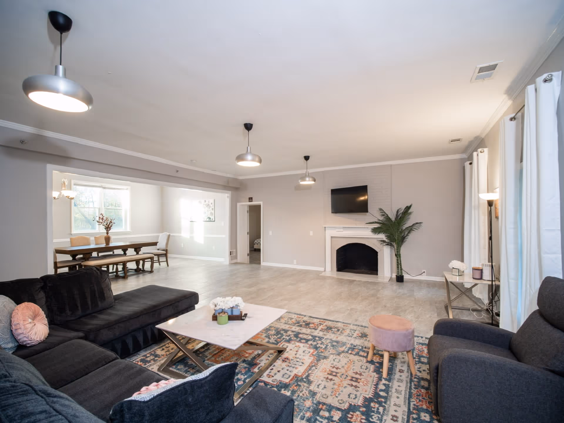 A spacious living room with a dark sectional sofa, a patterned area rug, a marble coffee table, and a small pink stool. The room features a white fireplace with a mounted flat-screen TV above it and a tall green plant beside it. There are three modern pendant lights hanging from the ceiling. In the background, there is a dining area with a wooden table and chairs near a window with white curtains.