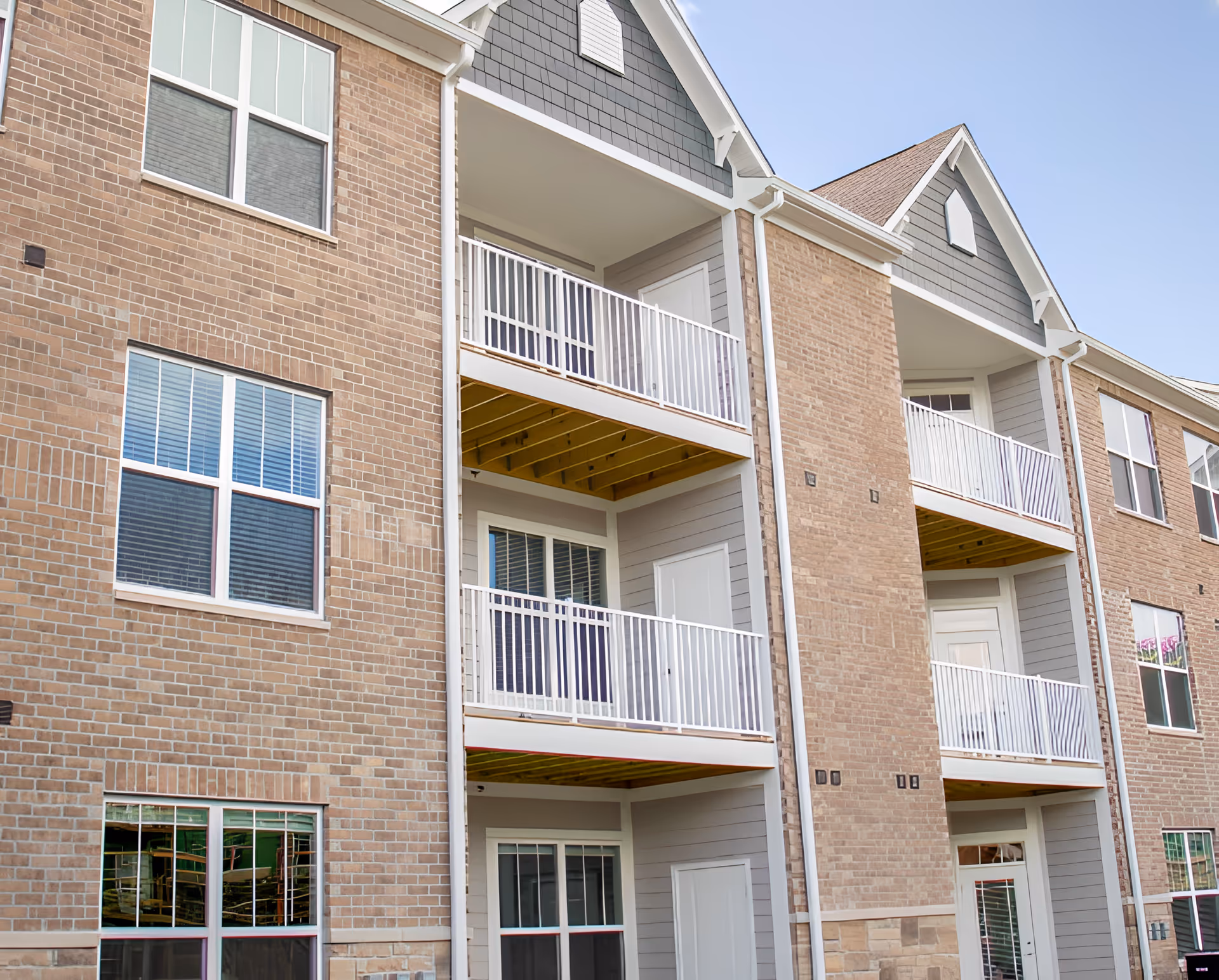 Exterior view of a multi-story brick and siding apartment building with balconies and white railings under a clear blue sky.