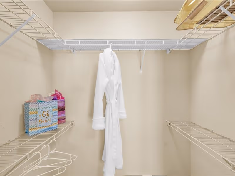 Interior view of a walk-in closet with beige walls and white wire shelving. A white bathrobe is hanging in the center, and there are two gift bags on the left shelf.