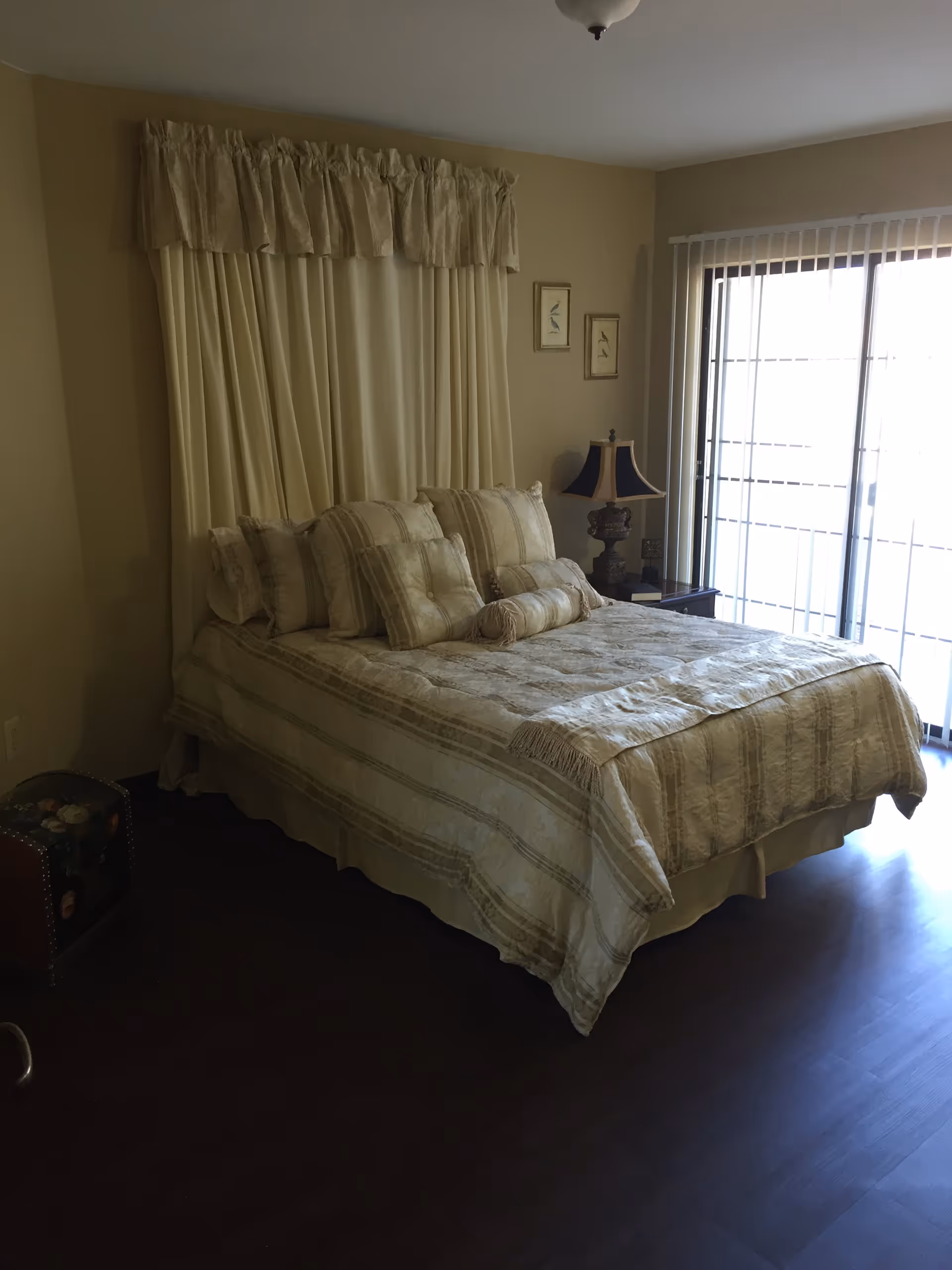 Bedroom with a neatly made bed with neutral linens, a bedside table and lamp, curtains, and a sliding glass door with vertical blinds.