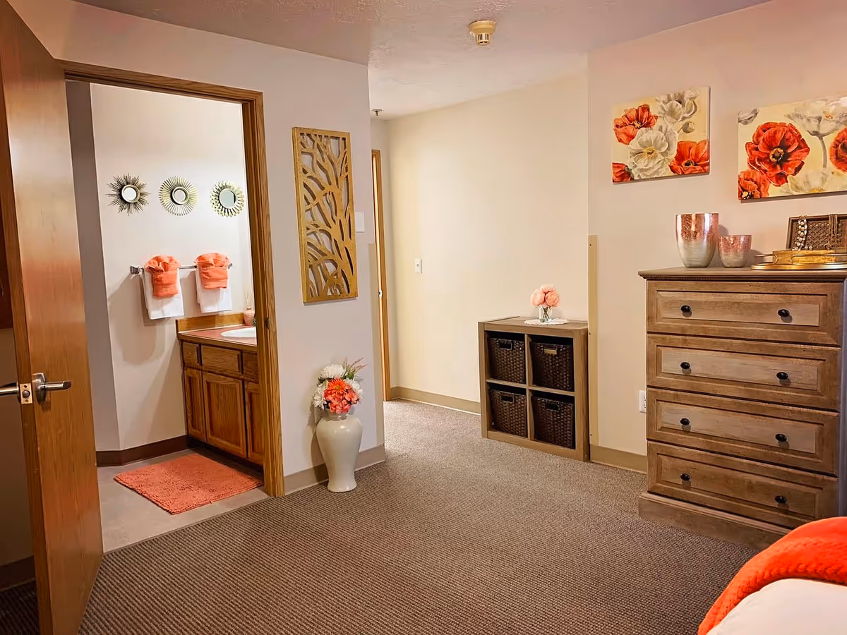 Interior view of a senior living facility room showing a wooden dresser with decorative items on top, a small shelving unit with baskets, floral wall art, and a doorway leading to a bathroom with a sink, mirror, and orange towels.