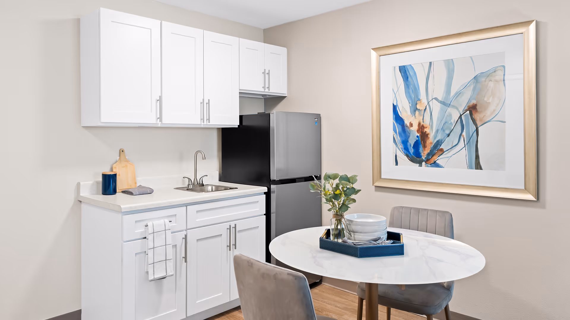 A small kitchen area with white cabinets, a stainless steel sink, and a silver refrigerator. Next to the kitchen is a round white table with two gray chairs. On the table is a blue tray holding white bowls and a small vase with green foliage. A large framed abstract painting with blue and brown tones hangs on the beige wall behind the table.