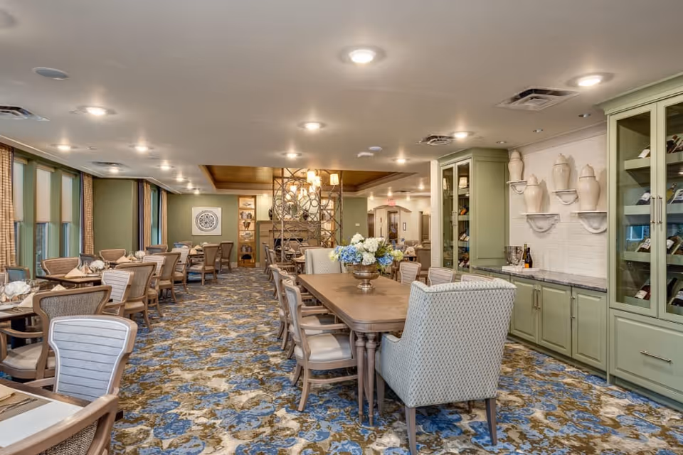A spacious dining room with multiple tables and chairs arranged neatly. The room features a patterned carpet in blue, beige, and brown tones, green cabinetry with decorative vases, and a large wooden dining table with a floral centerpiece. The walls are painted green and there are large windows with curtains along one side.