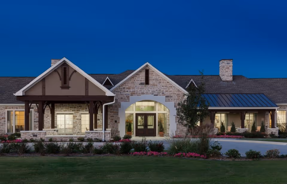 Exterior front view of a single-story stone building with a peaked roof and covered porch areas, illuminated at dusk with landscaped greenery and flowers in the foreground.