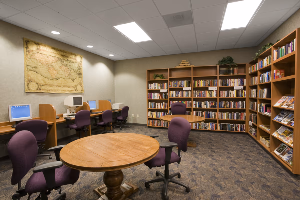 A quiet library or study room with wooden bookshelves filled with books and magazines. There are two round wooden tables with purple office chairs around them. Along one wall, there are computer workstations with purple chairs and desktop computers. A large vintage world map hangs on the wall above the computers. The room has a carpeted floor and a drop ceiling with fluorescent lighting.