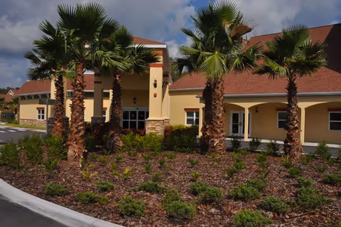 Exterior view of a senior living facility building with a covered entrance, surrounded by landscaped garden beds with palm trees and shrubs under a partly cloudy sky.