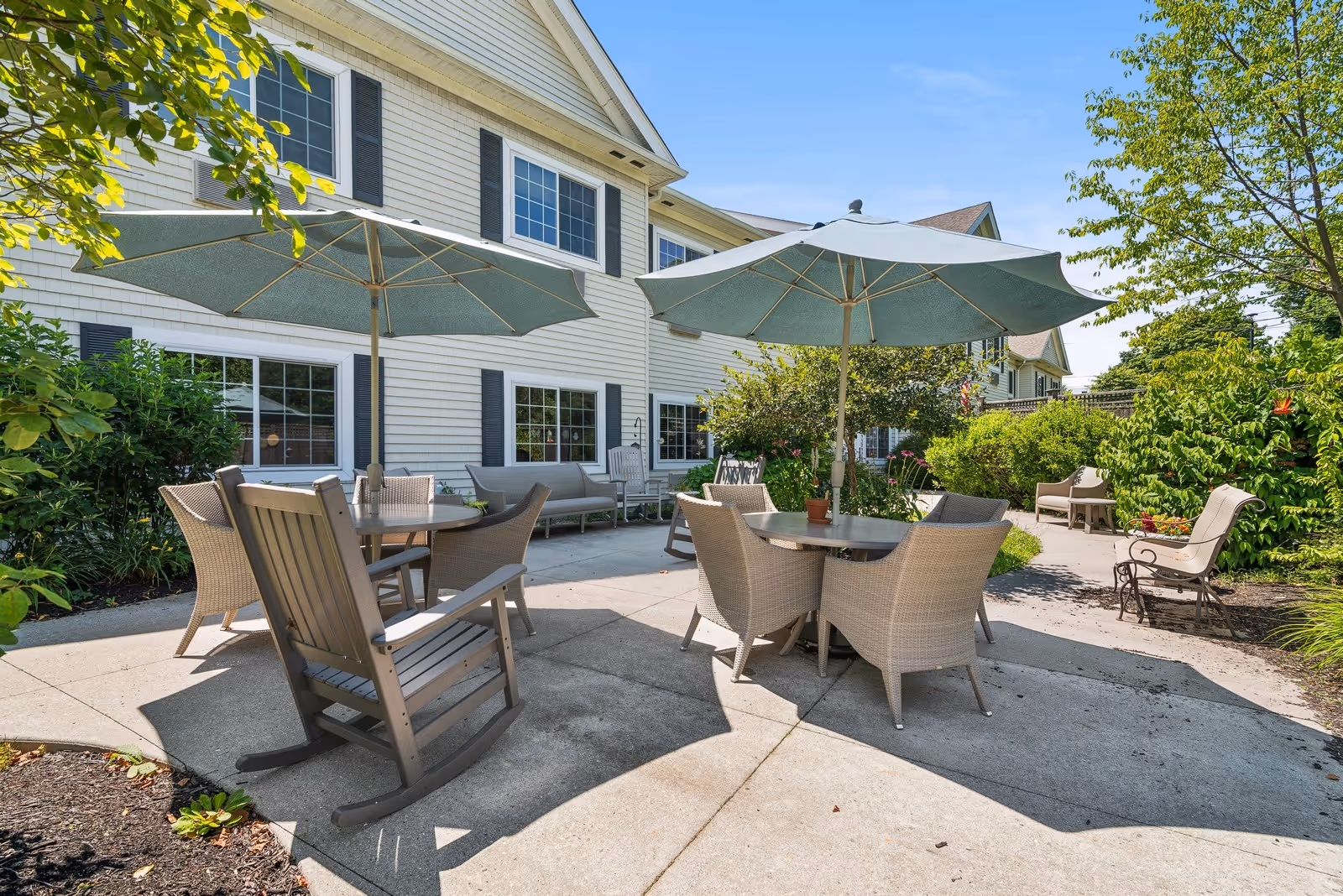 Outdoor patio area at The Enclave of Scarborough with round tables, wicker chairs, and large umbrellas providing shade. The patio is surrounded by greenery and shrubs, with a beige building featuring multiple windows in the background under a clear blue sky.
