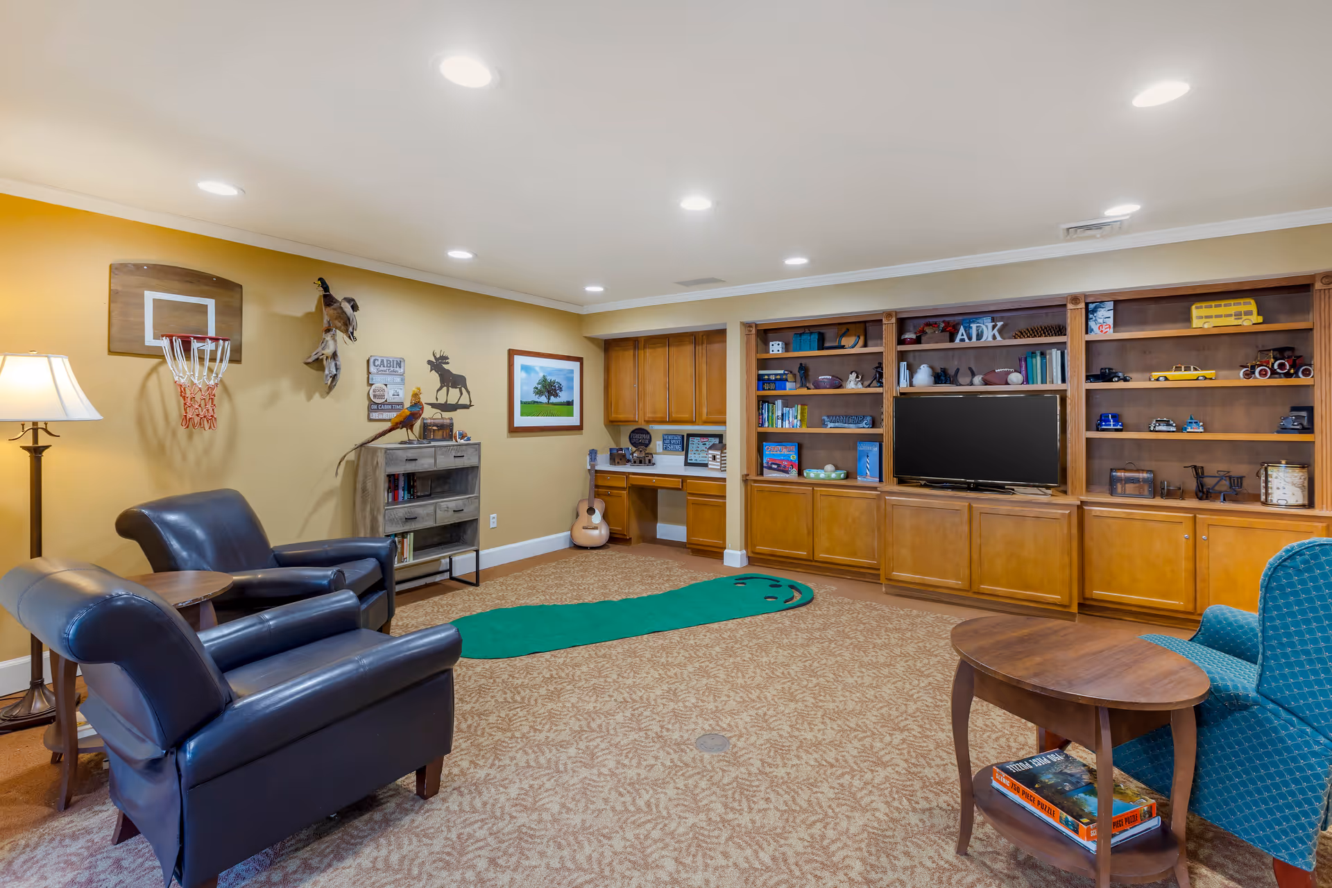 A cozy living room with beige walls and carpeted floor featuring two black leather armchairs, a blue upholstered chair, a wooden coffee table with books underneath, a small basketball hoop on the wall, a putting green mat on the floor, built-in wooden shelves filled with books and decorative items, a flat-screen TV, and a guitar resting against the wall.