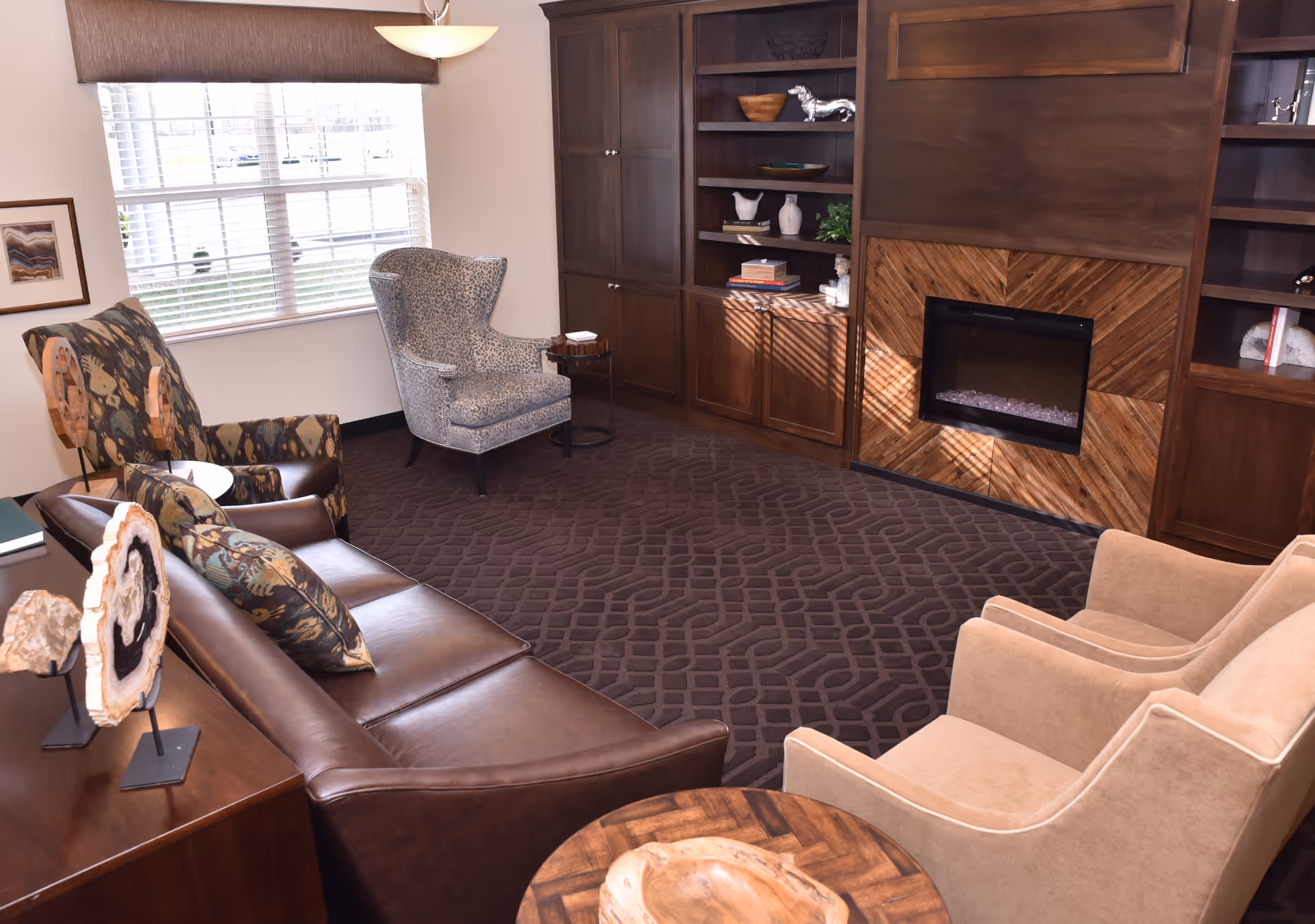 A cozy living room with a brown leather sofa, two beige armchairs, a patterned armchair near a window with blinds, a wooden coffee table, and built-in wooden shelves surrounding a modern electric fireplace.