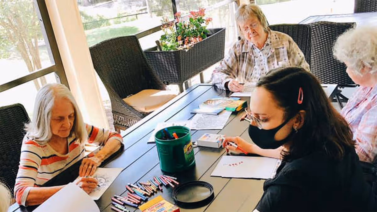 Four elderly women sitting around a rectangular table on a covered outdoor patio, engaged in coloring and drawing activities with colored pencils and paper. One woman is wearing a black face mask. The area is furnished with wicker chairs and has a planter with flowers in the background.