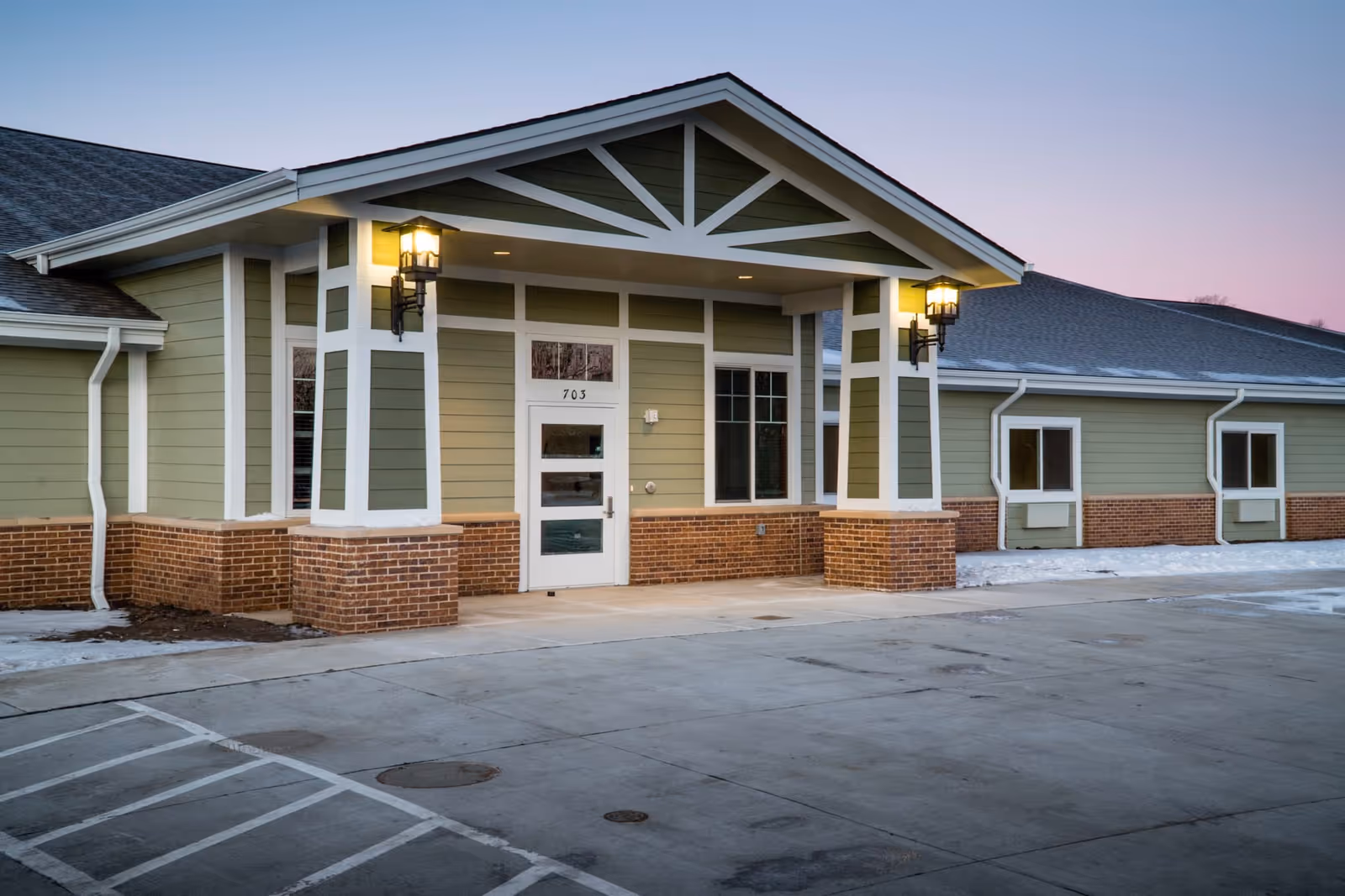 Exterior view of Hansen House Harlan building entrance with green siding, brick accents, two lit wall lanterns, and a covered porch area at dusk.