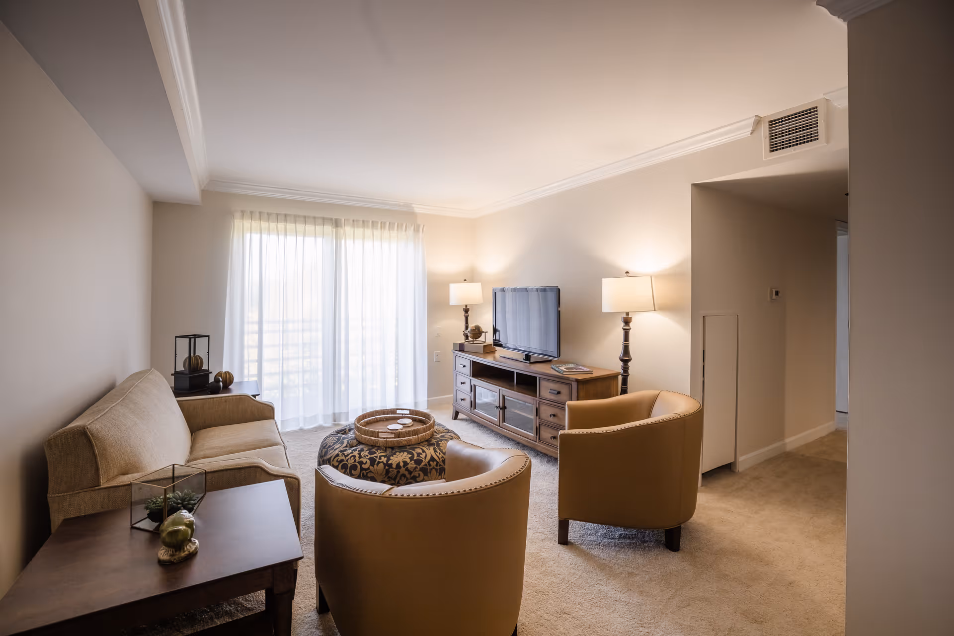 A cozy living room with beige walls and carpet, featuring a beige sofa, two tan armchairs, a round patterned ottoman, a wooden TV stand with a flat-screen television, two table lamps, and a side table with decorative items. Sheer curtains cover a large window letting in natural light.
