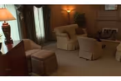 A cozy living room with beige upholstered armchairs and a sofa arranged around a coffee table. The room features wood-paneled walls, a floor lamp, a table lamp on a wooden dresser, and a window with sheer curtains allowing soft natural light.