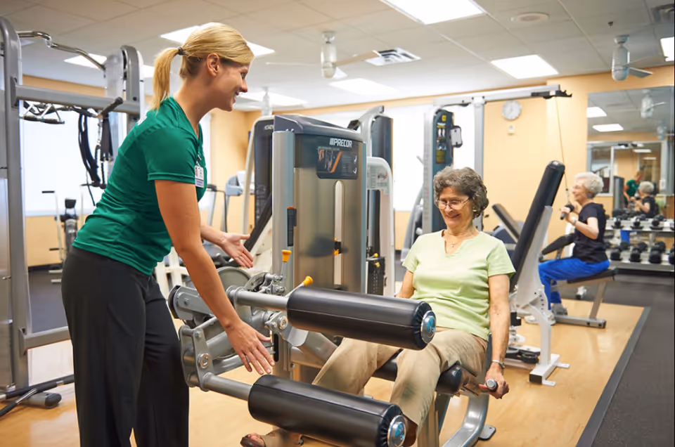An instructor assists a seated woman using a leg-extension machine in a fitness room while another woman works out in the background.