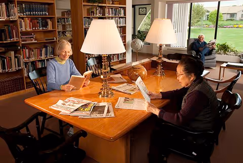 Two elderly women sitting at a wooden table in a library or reading room, reading books and newspapers under two table lamps. Bookshelves filled with books are visible in the background. A man is sitting on a chair near a large window, reading a newspaper, with a view of a green lawn and houses outside.