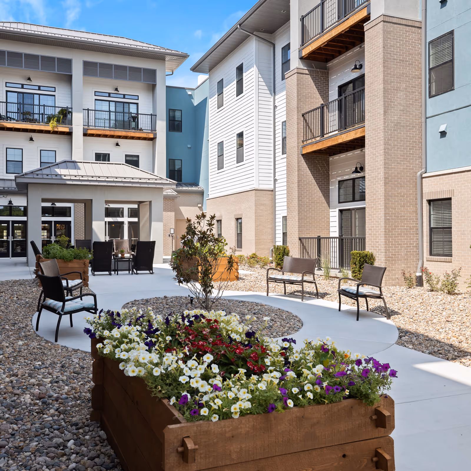Outdoor courtyard area of a senior living facility with a curved concrete walkway, several chairs with cushions, wooden planter boxes filled with colorful flowers, and multi-story buildings with balconies surrounding the space under a clear blue sky.
