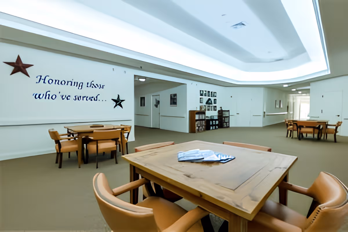 A spacious common area with multiple wooden tables and chairs arranged for seating. The walls are white with a decorative message that reads 'Honoring those who've served...' accompanied by two star decorations. The ceiling features recessed lighting with a modern design. Shelving units and framed pictures are visible along the far wall.