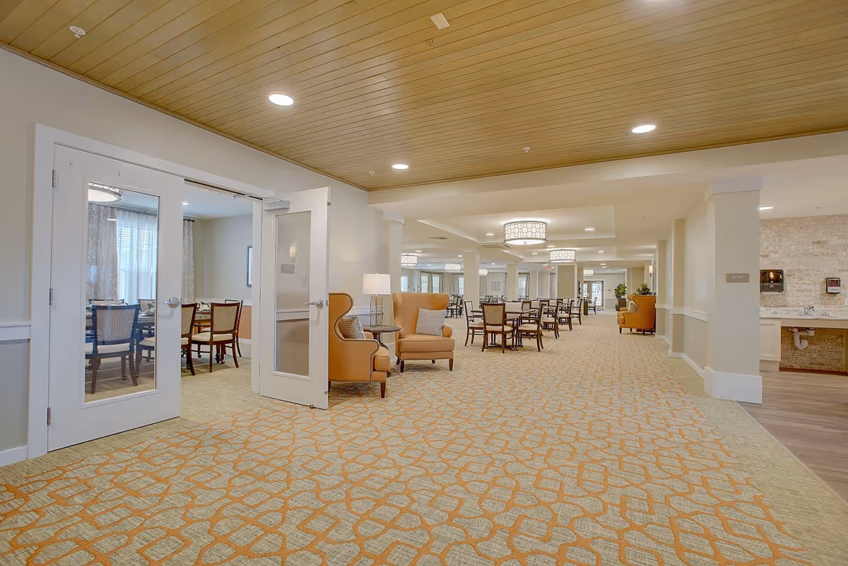 Spacious and well-lit interior of a senior living facility featuring a large carpeted hallway with patterned flooring, several round tables with chairs arranged for dining, and comfortable armchairs with side tables and lamps. There is a room with glass double doors on the left containing a dining table and chairs, and a sink area on the right side of the hallway. The ceiling has recessed lighting and decorative light fixtures.