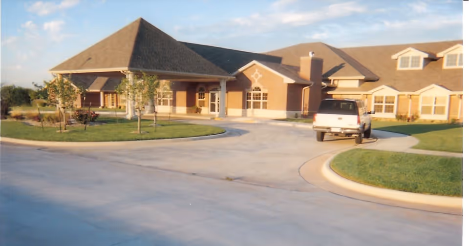 Exterior view of a single-story brick building with a large covered entrance and multiple windows, surrounded by a circular driveway and landscaped with grass and small trees. A white SUV is parked near the entrance.