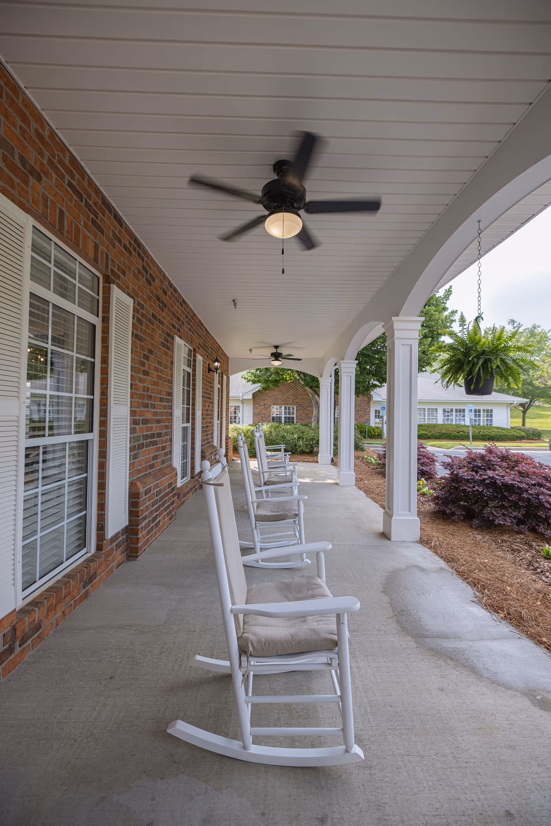 Covered outdoor porch area with white rocking chairs lined up along a brick wall with windows. Ceiling fans with lights hang from the white ceiling. There are white columns supporting the porch roof and hanging plants. Shrubs and greenery are visible beyond the porch.