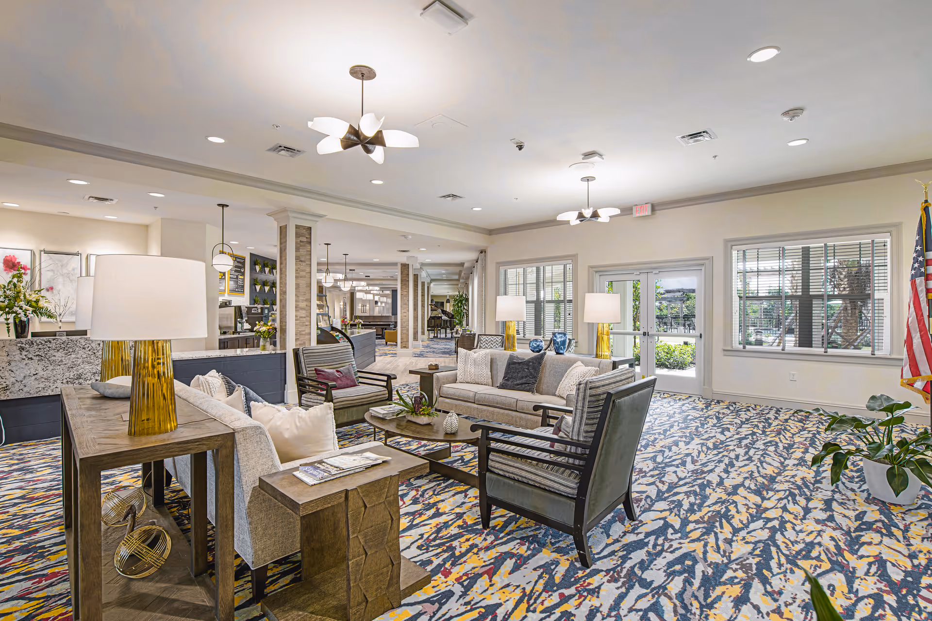 A spacious and well-lit living room area in a senior living facility with comfortable sofas and armchairs arranged around a coffee table. The room features modern light fixtures, large windows with blinds, decorative plants, and an American flag near the entrance door. The carpet has a colorful abstract pattern, and the overall decor is warm and inviting.