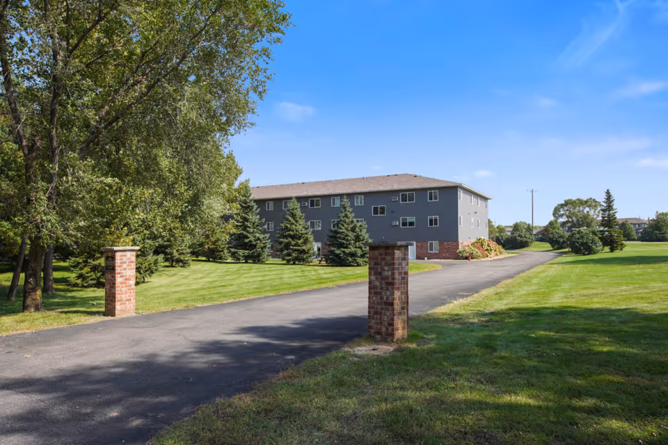 A paved driveway flanked by two brick pillars leads to a three-story gray building surrounded by green grass, trees, and shrubs under a clear blue sky.