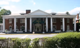 Front façade of a two-story brick building with large white columns, a central entrance with a green awning, and hedges in the foreground.