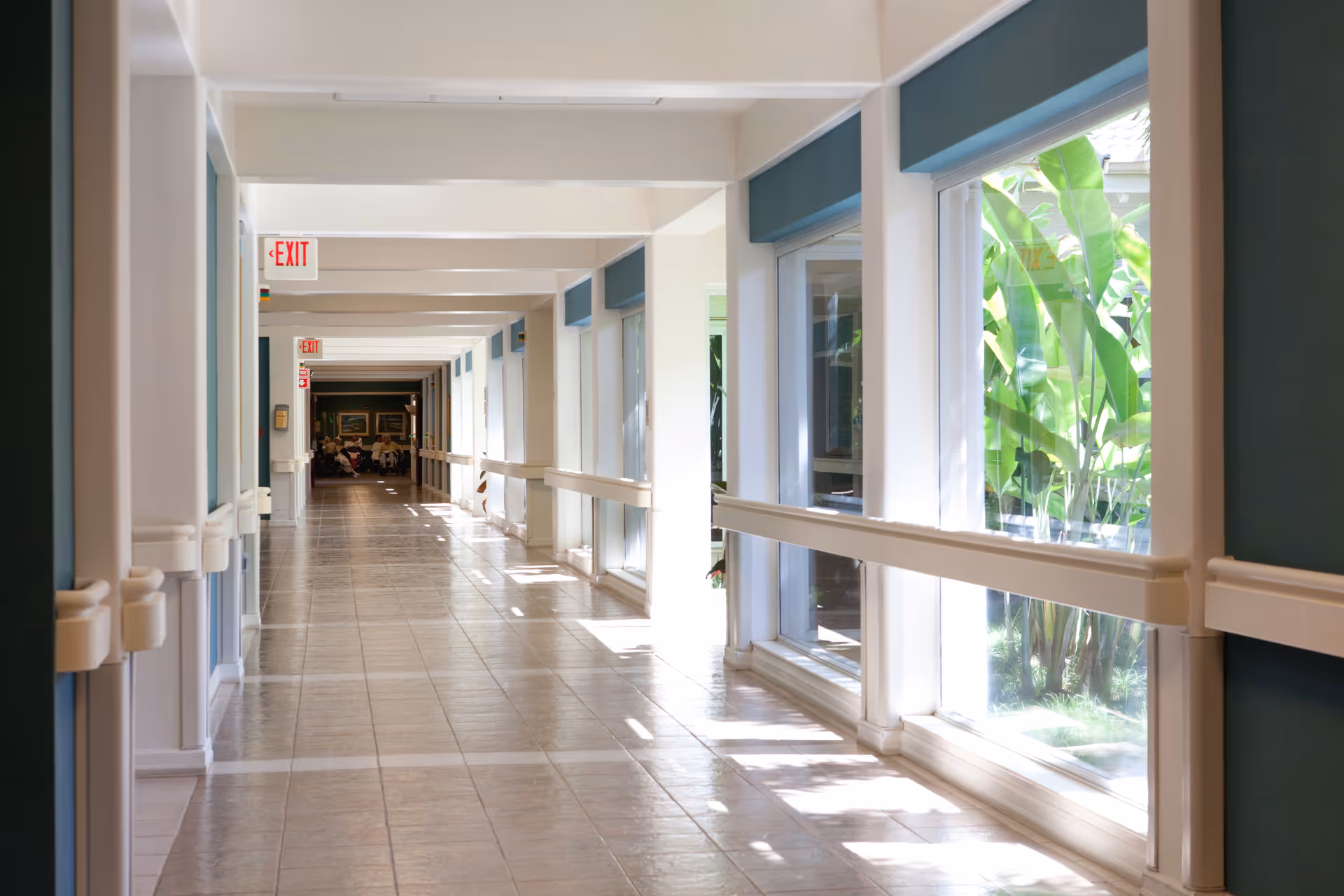 A long, bright hallway with large windows on the right side letting in natural light and showing green plants outside. The hallway has tiled floors, handrails along both walls, and exit signs hanging from the ceiling. At the far end, a group of people is seated.