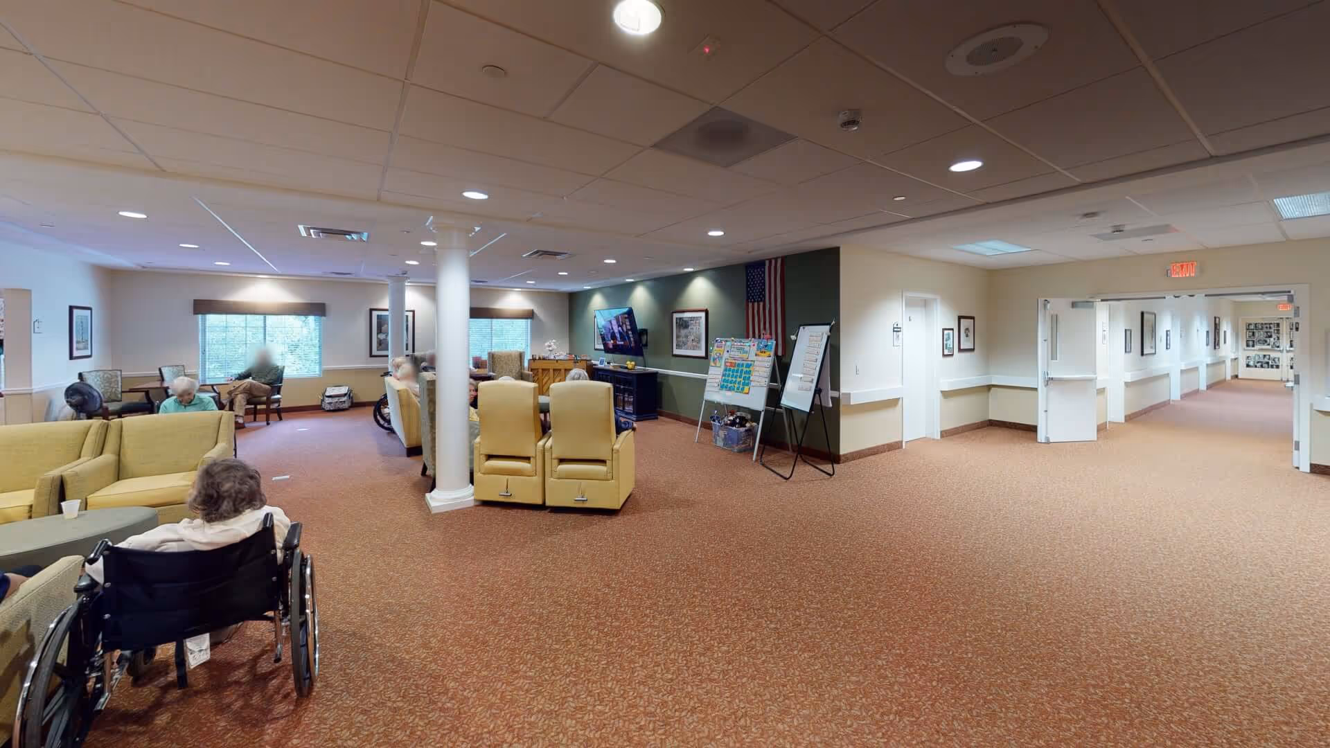A spacious common area in a senior living facility with several elderly residents seated in armchairs and wheelchairs. The room has carpeted floors, beige walls, and a green accent wall with an American flag, a TV mounted on the wall, and two whiteboards on stands. There are large windows letting in natural light and a hallway with double doors leading to other parts of the facility.
