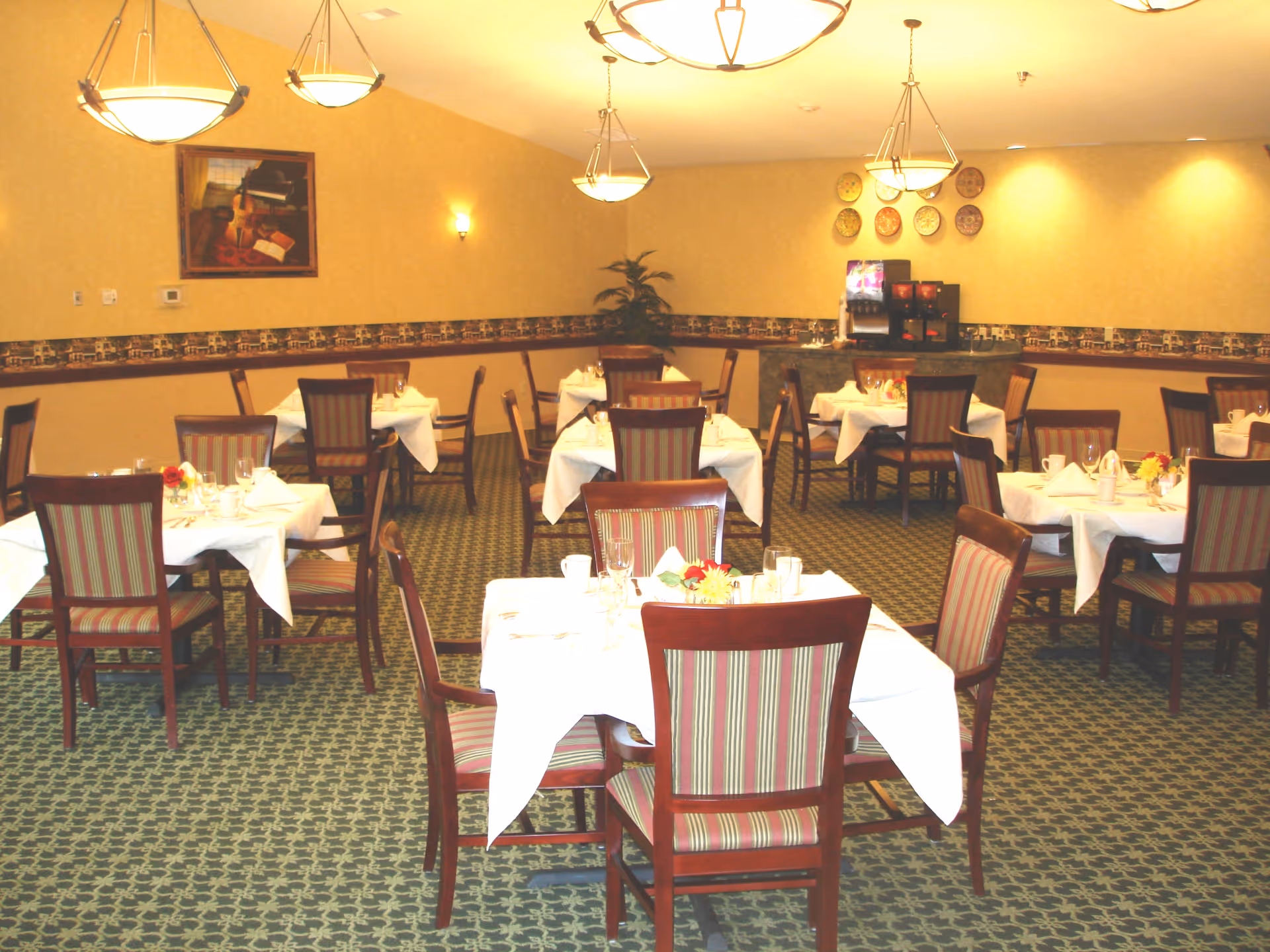 A dining room with multiple tables covered in white tablecloths, each set with glasses, cups, and small flower arrangements. The room has patterned carpet, beige walls with a decorative border, and several hanging light fixtures. There is a beverage station against the far wall and framed artwork on the wall.