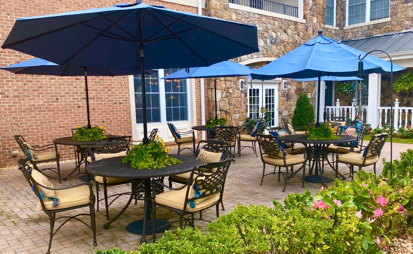 Outdoor patio area at The Woodlands Retirement Community featuring several round metal tables with beige cushioned chairs and blue umbrellas. The patio is paved with bricks and surrounded by greenery and flowering plants. The building exterior is made of brick and stone with windows and doors visible.