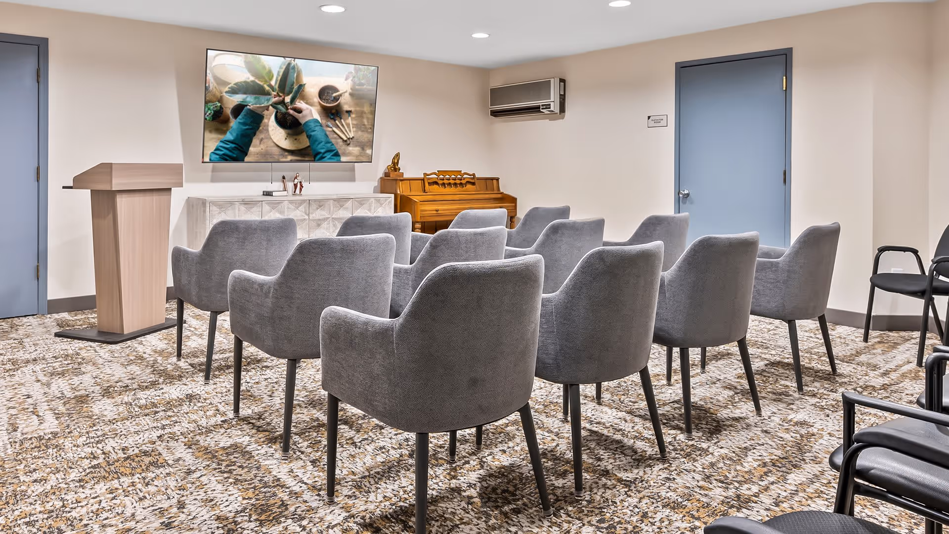 A small conference or presentation room with two rows of gray upholstered chairs facing a wooden podium and a large wall-mounted TV screen displaying a person planting a small plant. There is a wooden piano against the back wall, two blue doors, and a patterned carpet on the floor.