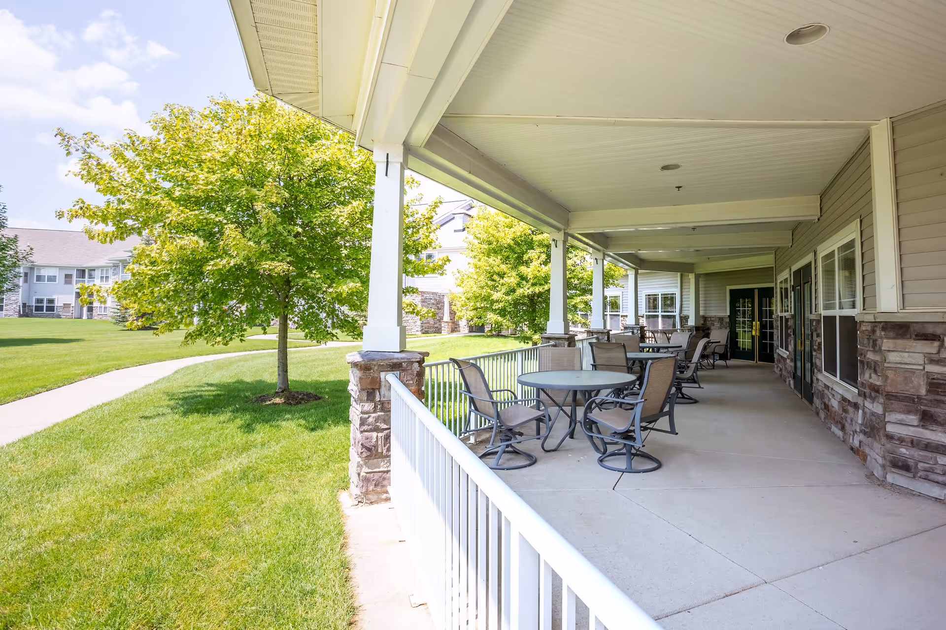 Covered outdoor patio area with several round tables and chairs, adjacent to a building with stone and siding exterior. Green grass, trees, and a paved walkway are visible in the background under a partly cloudy sky.