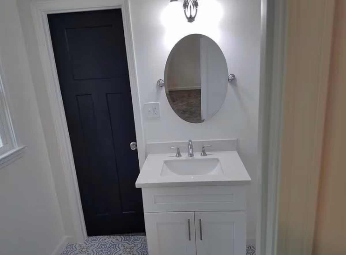 Bathroom vanity with a white sink and oval mirror, light fixture above, patterned tile floor, and a dark door to the left.