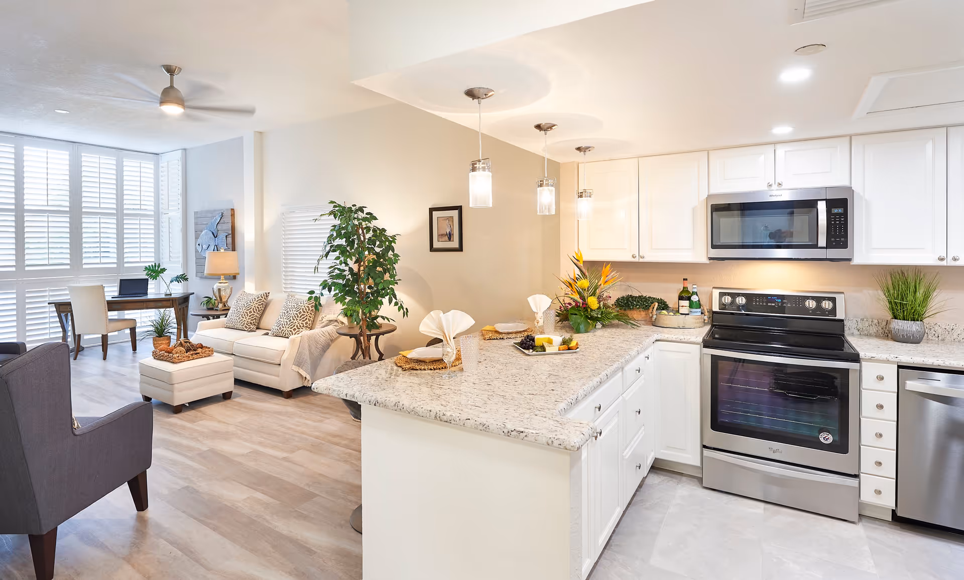 Bright and modern open-concept living space featuring a kitchen with white cabinets, granite countertops, stainless steel appliances including a stove, microwave, and dishwasher. The kitchen counter is set with plates, glasses, and a floral centerpiece. Adjacent to the kitchen is a cozy living area with a white sofa, patterned pillows, a small ottoman, a gray armchair, a wooden desk with a laptop, and large windows with white shutters allowing natural light to fill the room.