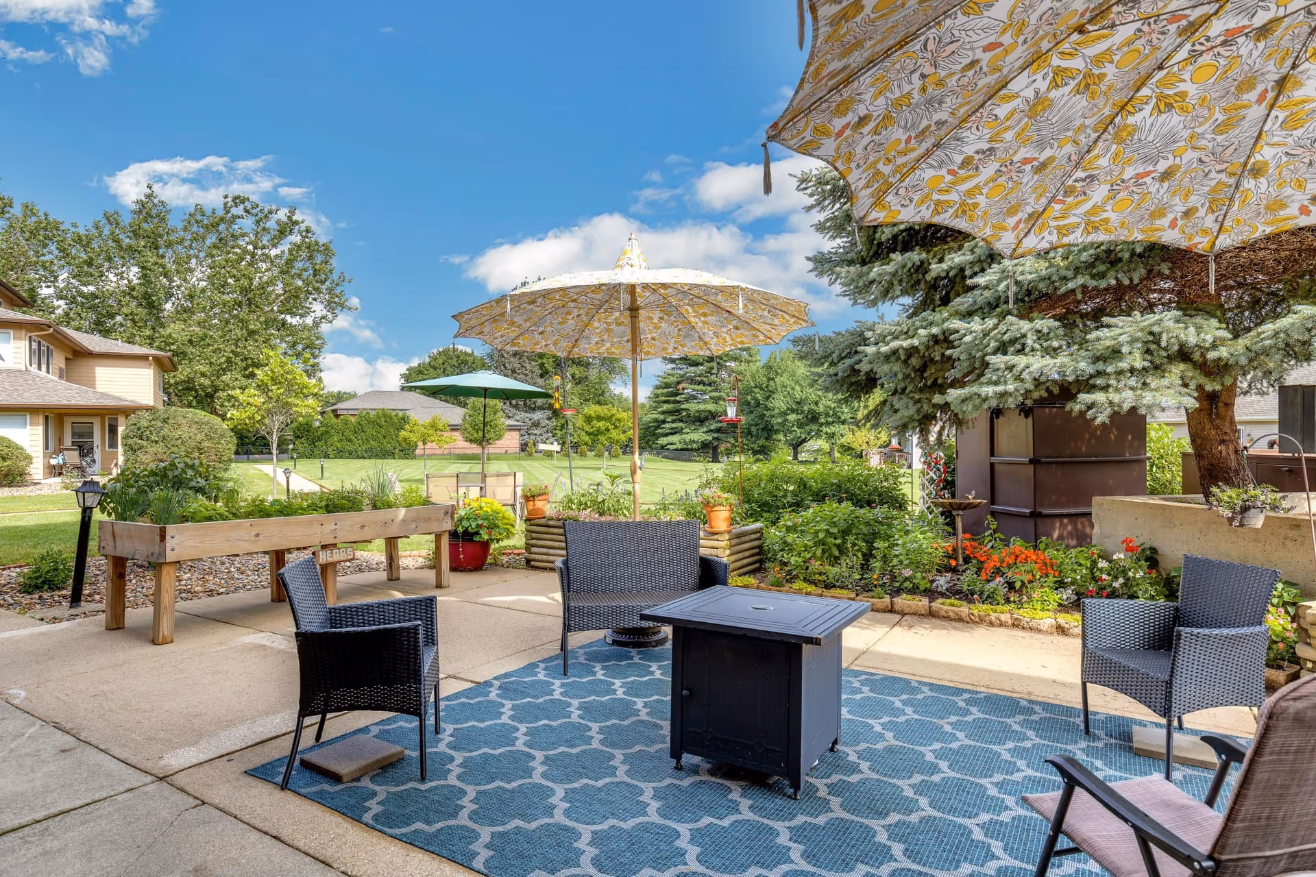 Outdoor patio with umbrellas, wicker chairs around a central table on a patterned rug, surrounded by planters and lawn.