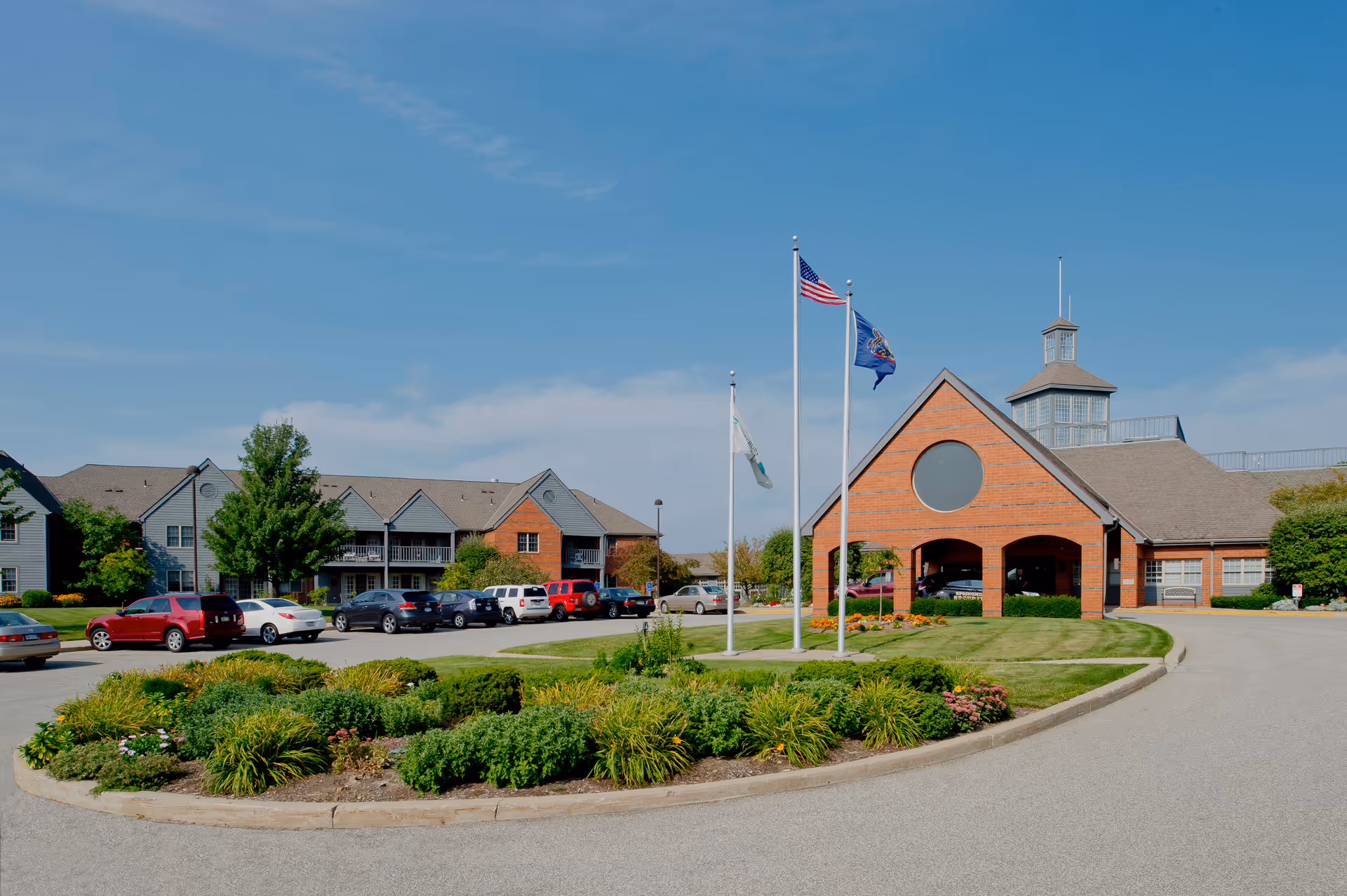 Exterior view of Springhill Senior Living facility showing a large brick and siding building with a peaked roof and a circular window above the entrance. Three flagpoles with flags stand in front of the building, surrounded by well-maintained landscaping and a circular driveway with parked cars.