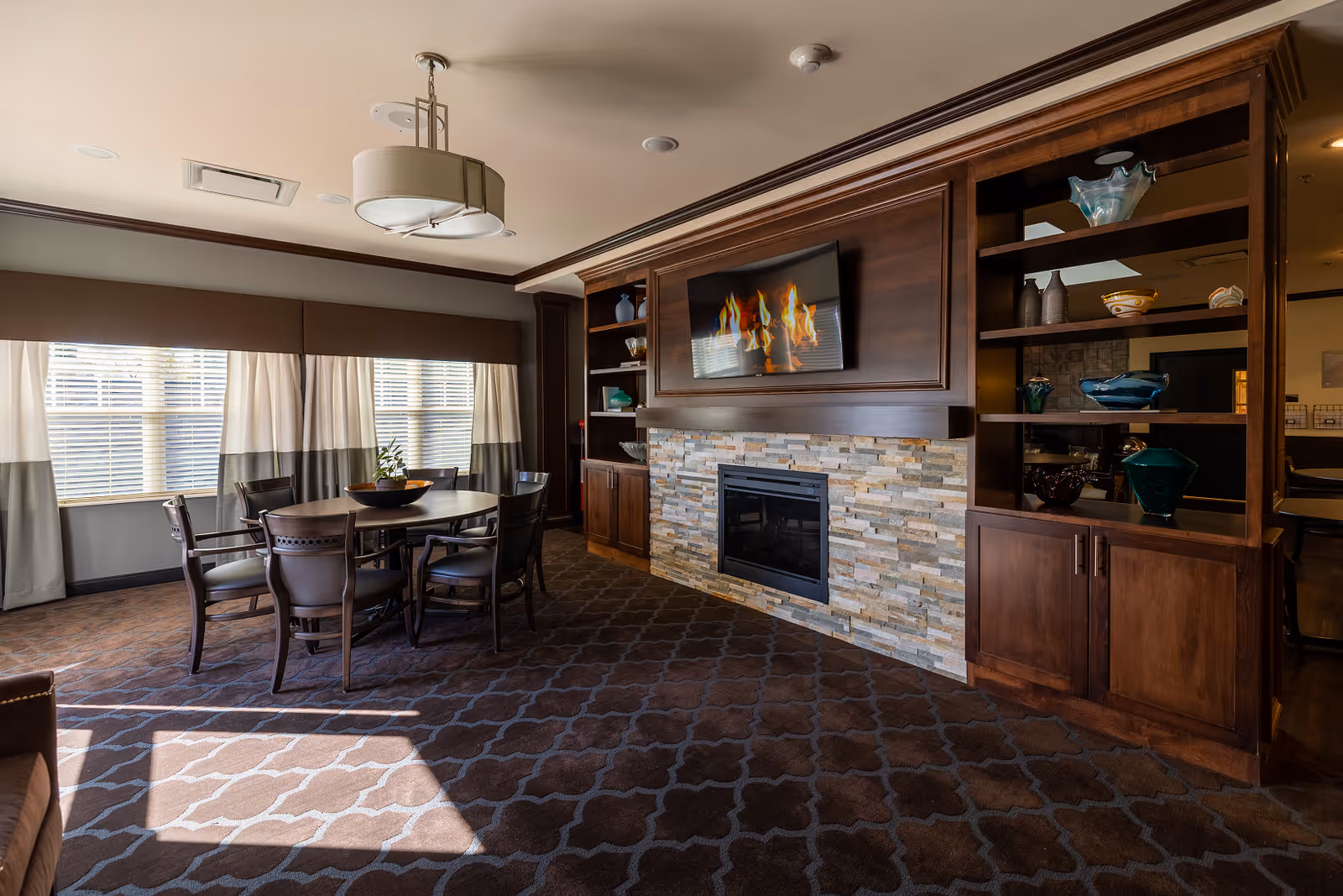 A warm common room with a round dining table and chairs, built-in shelving, and a stone-front fireplace with a mounted TV.