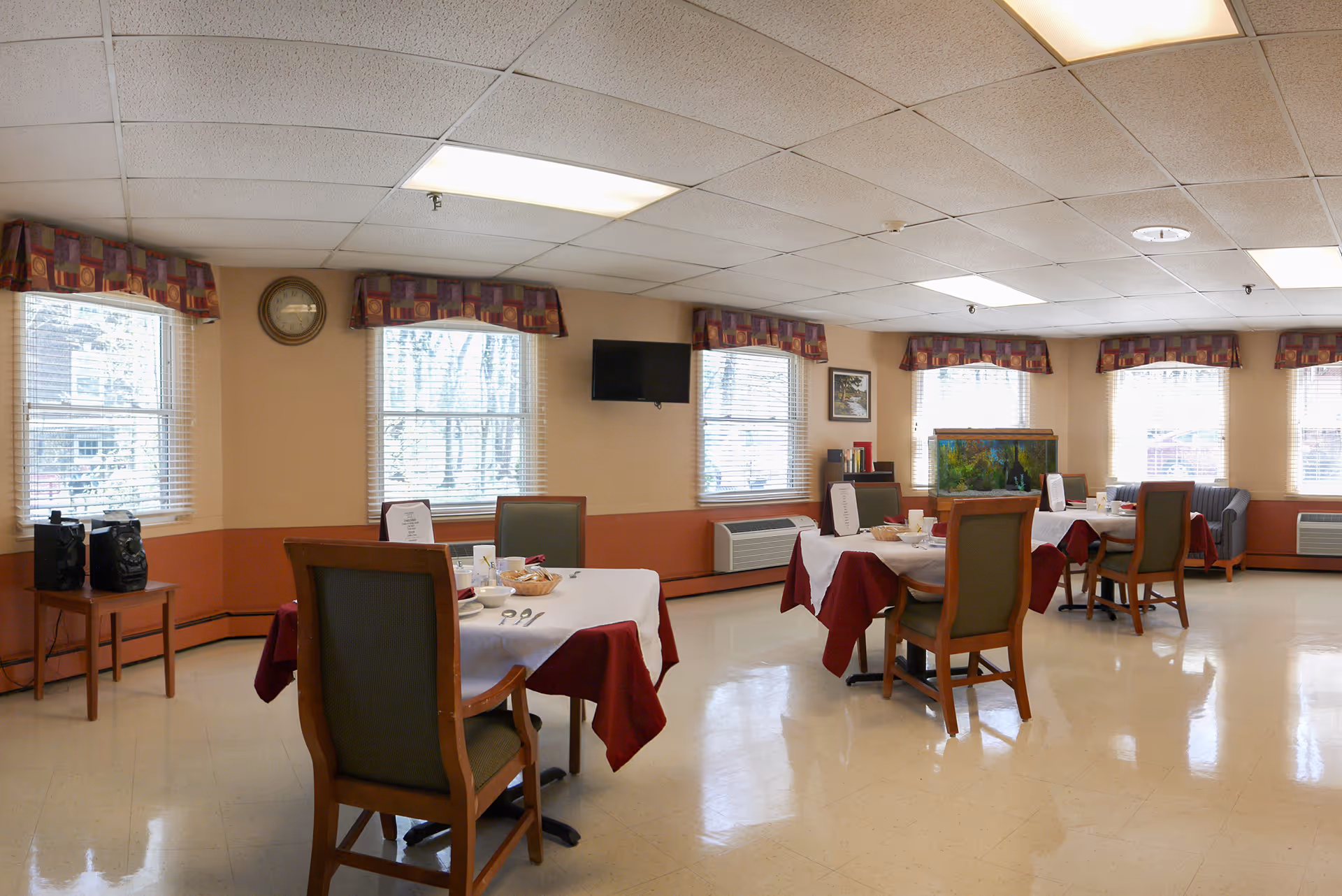 A dining room in Cedarwood Plaza with several tables covered in white and red tablecloths, each set with dishes, cups, and utensils. The room has multiple windows with patterned valances, a wall clock, a mounted TV, and an aquarium. There are chairs around the tables and a small table with a stereo system in the corner.