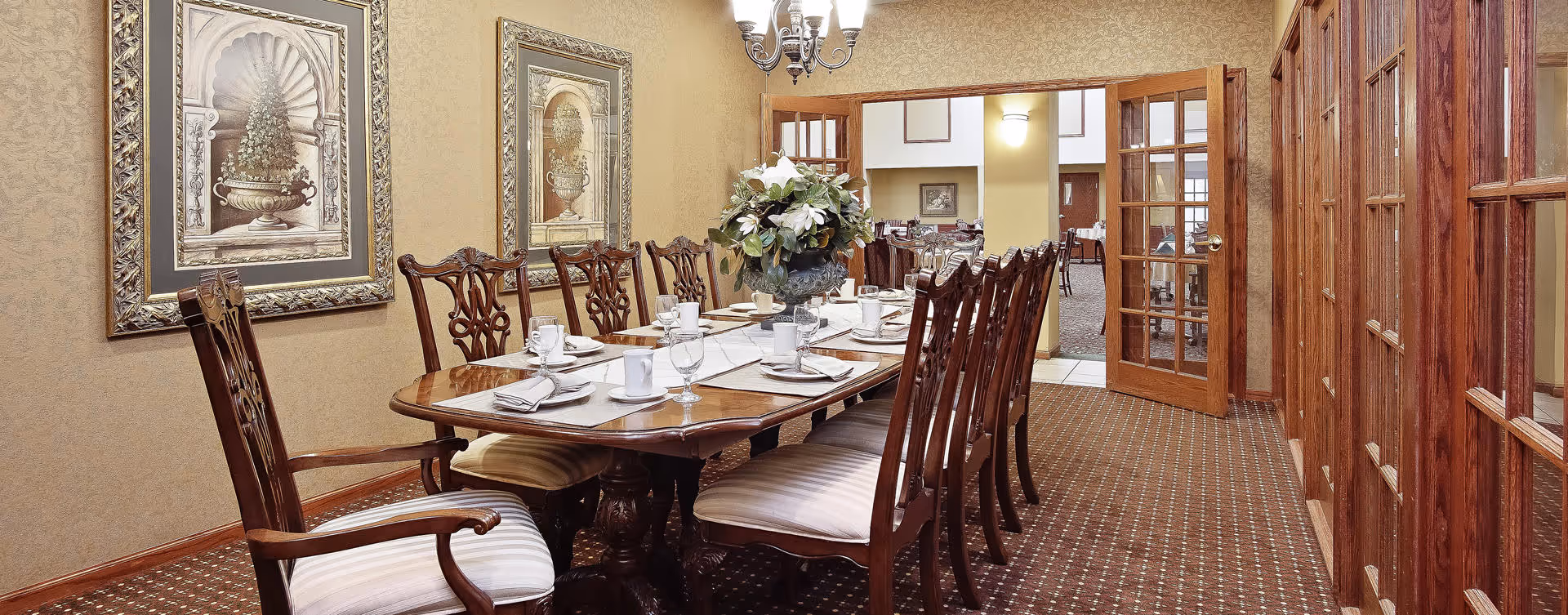 A formal dining room with a long wooden table set for a meal, surrounded by wooden chairs with cushioned seats. The walls are decorated with framed artwork, and a chandelier hangs above the table. There is a floral centerpiece on the table, and glass-paneled wooden doors lead to another room in the background.