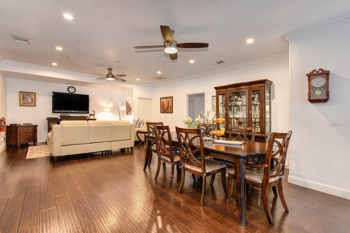 A spacious and well-lit living and dining area with wooden flooring. The dining area features a dark wooden table with eight matching chairs, a centerpiece with flowers, and a glass-fronted cabinet displaying dishes. The living area has a beige leather sofa facing a wall-mounted flat-screen TV, with a wooden side table and a floor lamp nearby. The ceiling has recessed lighting and two ceiling fans. The walls are decorated with framed pictures and a wall clock.