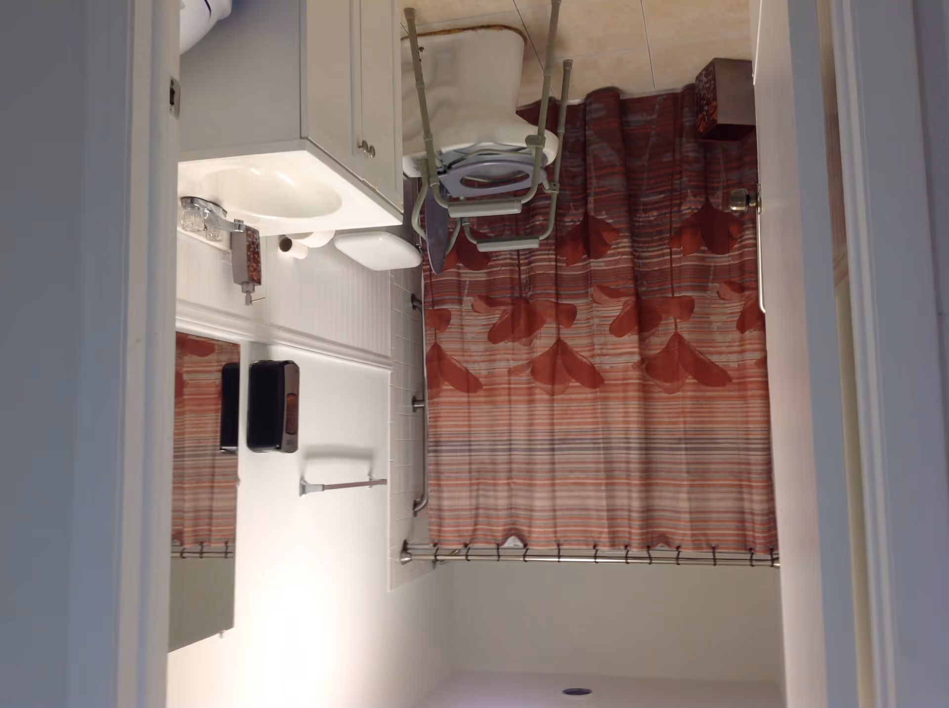 A bathroom with a white sink and cabinet, a toilet, a shower with a red and orange patterned curtain, and a white chair with armrests placed inside the shower area.