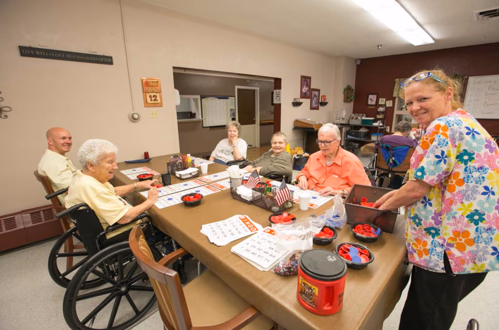 A group of elderly people and a caregiver gathered around a table playing bingo in a common room. The table is covered with bingo cards, red and blue bingo chips, and small American flags. The caregiver is smiling and holding a container of bingo chips. The room has beige walls with some decorations and a sign that reads 'LIVE WELL • LOVE MUCH • LAUGH OFTEN'.
