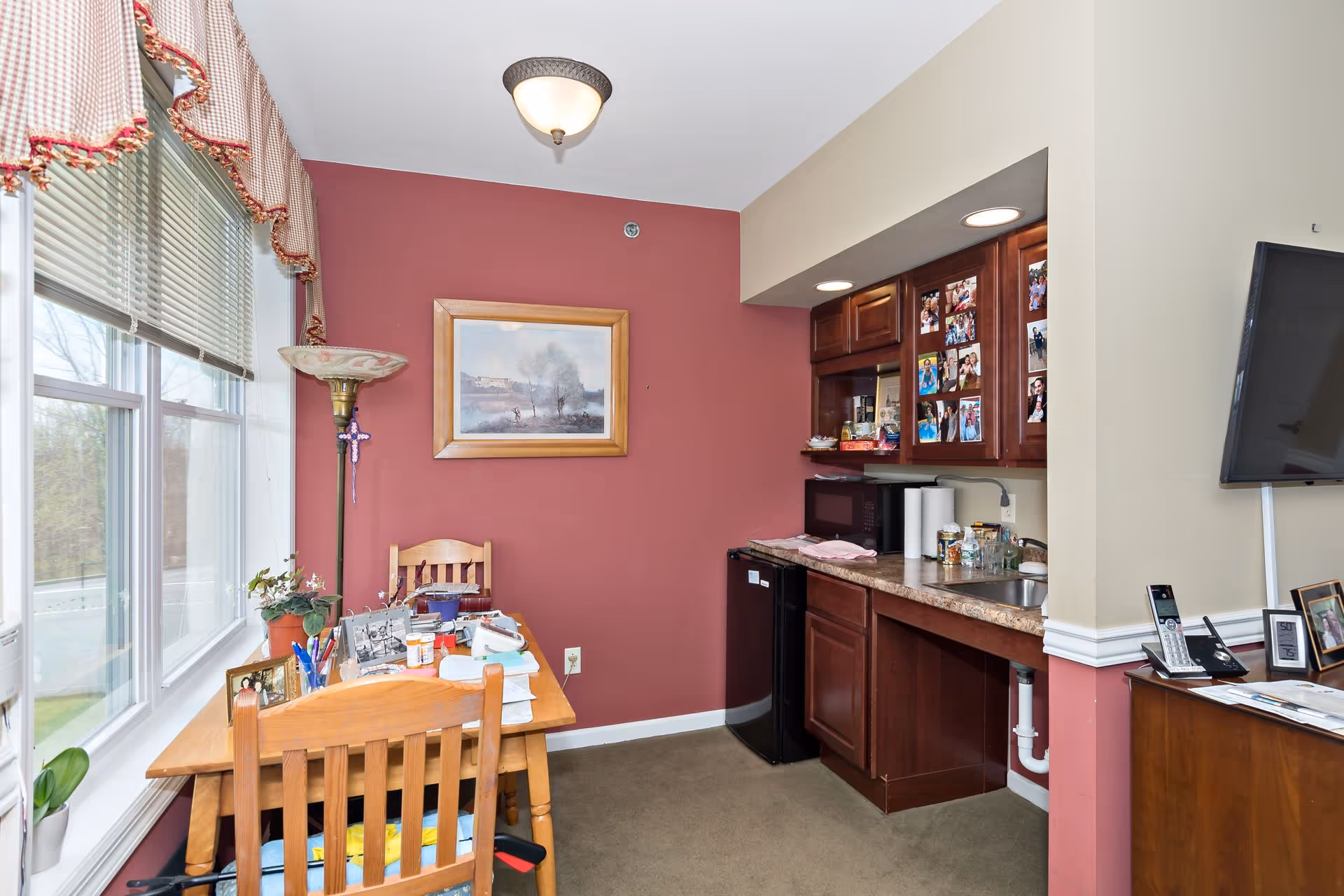 A small dining and kitchenette area with a wooden table and chairs near a window with blinds and a checkered valance. The walls are painted in two tones, with one wall in a deep red color and the others in beige. The kitchenette has dark wood cabinets, a countertop with a sink, a microwave, and a mini fridge. Family photos are displayed on the cabinet doors. A floor lamp stands next to the table, and a framed painting hangs on the red wall.