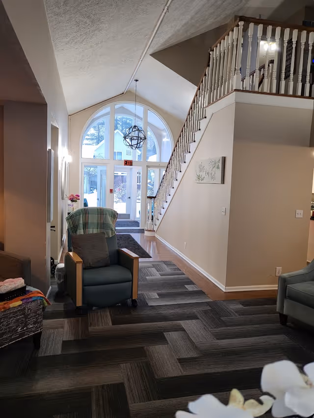 Lobby of a senior living facility with seating, patterned carpet, a staircase to an upper level, and large arched glass entrance doors.