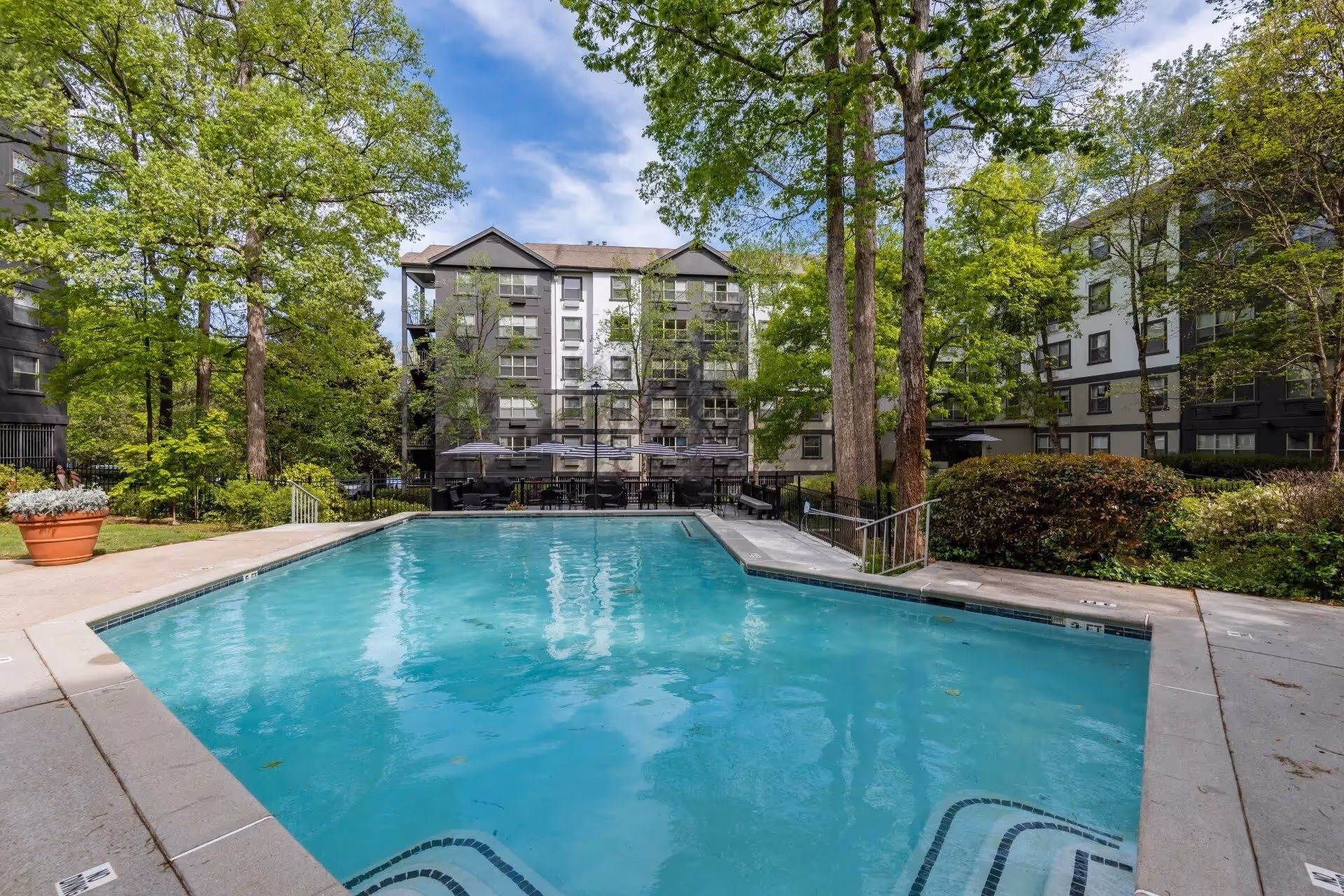 Outdoor swimming pool surrounded by concrete deck with lounge chairs and umbrellas, set in a landscaped area with trees and bushes, with multi-story residential buildings in the background under a partly cloudy sky.